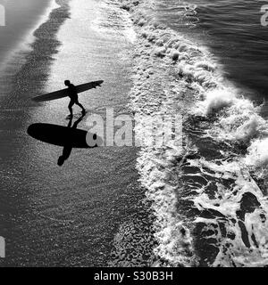 Surfista maschio passeggiate fino alla spiaggia dopo la navigazione. Manhattan Beach, California, Stati Uniti d'America. Foto Stock