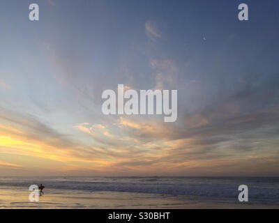 Un surfista lascia il oceano Atlantico dopo il tramonto, Kommetjie Long Beach, Cape Town, Sud Africa. 2019. Foto Stock