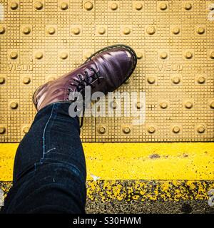 Un piede in un Dr Martens boot sta a cavallo tra il giallo "stare dietro la linea di marcatura" su un regno unito piattaforma ferroviaria Foto Stock