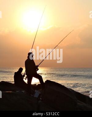 Una coppia di pescatori stagliano dal sole che si alza in piedi sulle rocce al mare. Foto Stock