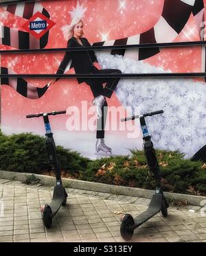 Due scooter elettrici disponibili per noleggio parcheggiato nella pavimentazione di fronte a un muro vicino a una stazione della metropolitana, essendo la parete dipinta con un design murale Foto Stock