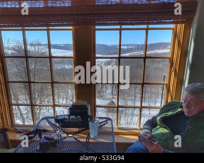 Dubuque, IOWA, 23/02/2020–Closeup foto di uomo controllare il suo telefono mentre pranzo di fronte a una panata finestra con bella vista del paesaggio di terreno agricolo coperto di neve in giornata di sole. Foto Stock