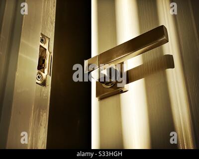 La vista a primo piano di una moderna maniglia della porta cromata e le ombre dalle luci geometriche e dalle strisce scure del sole risplendono attraverso le tende veneziane Foto Stock