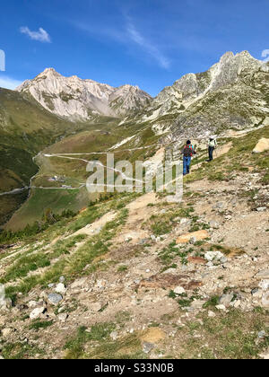 Due uomini con zaini e bastoni a piedi che percorrano il ripido sentiero della Via Francigena nelle Alpi Italiane. Bel cielo blu, roccioso terreno zigzag. Foto Stock