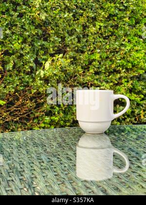 Tazza bianca per caffè su mobili da giardino con piano in vetro riflettente Foto Stock