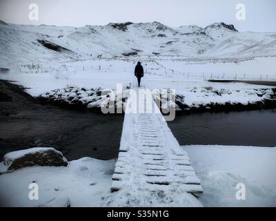 Isolamento. Una figura scura si erge con la schiena alla telecamera sulla fine di un ponte innevato su un fiume. Un paesaggio montano innevato sullo sfondo. Foto Stock