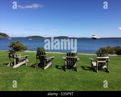 Paesaggio con quattro sedie Adirondack su un prato che si affaccia sull'oceano e una nave da crociera in lontananza Foto Stock