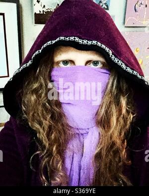 Donna dai capelli rossi con occhi blu che indossa una felpa con cappuccio viola e una sciarpa viola come maschera COVID19 che copre il viso. Foto Stock