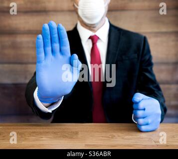 Un medico o un uomo d'affari di Coronavirus che indossa dpi, compresi i guanti chirurgici blu e la maschera facciale che tiene la mano in su in un gesto di arresto per mantenere le persone di distanza sociale e auto-isolamento Foto Stock