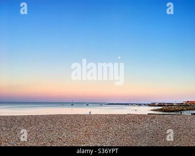 Tramonto che si raduna su un mare calmo con una superficie simile a uno specchio. Blu intenso, giallo e viola nel cielo, e una piccola luna. Spiaggia di ciottoli con piccole figure. Piccole imbarcazioni appena fuori al mare. Case lontane. Foto Stock