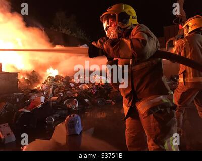 Un pompiere tiene un ramo e un tubo flessibile mentre spruzzano acqua su un incendio in un impianto di riciclaggio. Foto Stock