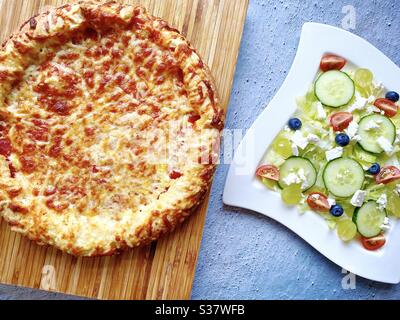 Vista dall'alto del formaggio in padella e della pizza al pomodoro appena sfornati con due piatti di insalata feta fresca. Pizza cheesy con crosta spessa contro un bancone di cucina di cemento grezzo. Fotografia di cibo piatto. Foto Stock