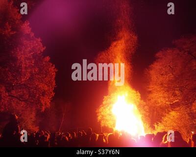 Una fotografia di una folla si trovava di fronte ad un grande falò in un bosco nella notte del falò. Grandi fiamme e scintille che raggiungono il cielo notturno. Foto Stock