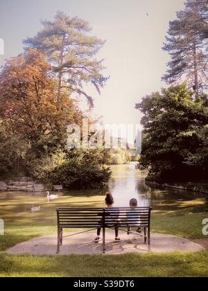 Misto ragazza e ragazzo da corsa sedette su panchina con vista lago con cigni a Bedford Park, Bedfordshire, Inghilterra, Regno Unito. Foto Stock