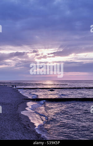 Costa al largo di Kuehlungsborn sull'isola tedesca Ruegen nel mistica serata viola con alcuni passeggini sulla spiaggia di sabbia spiaggia con groynes che sporgono nell'acqua Foto Stock