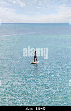 Un SUP Paddler in piedi con i tronchi blu per nuotare il mare aperto sulle acque turchesi blu e sotto un cielo leggermente nuvoloso Foto Stock