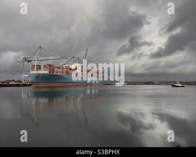 A tug boat (right) sails along Southampton Water past a container ship being unloaded at Southampton Docks in England. Stock Photo