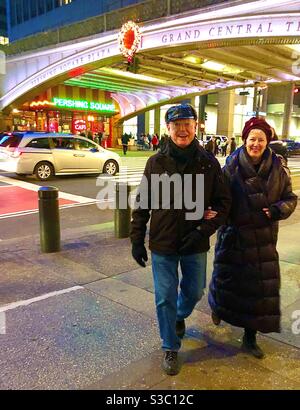 Vestiti con abiti caldi e cappelli in una fredda notte invernale, un uomo e una donna passeggiano allegramente al braccio da Grand Central sulla 42nd Street a New York City, USA. Foto Stock