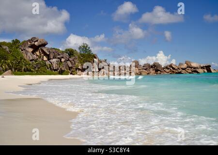 Belle onde turchesi tropicali dell'Oceano Indiano lambiscono delicatamente lungo il spiaggia di sabbia bianca circondata da grandi rocce levigate su un Giornata luminosa e soleggiata nelle Seychelles Foto Stock