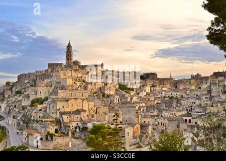 La più magica e bella città collinare antica di Matera Nel sud Italia al tramonto con un cielo morbido opalescente e luce dorata che splende sulle case e sulla chiesa sottostante Foto Stock