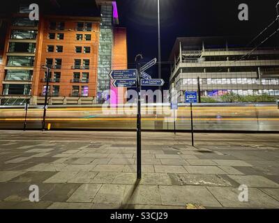 Lunga esposizione di un tram nel centro di Manchester City con edifici moderni sullo sfondo. Foto notturna con sentieri luminosi. Foto Stock