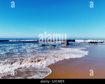 Onde che si lavano sulla spiaggia sabbiosa tra le rocce in una giornata di sole limpida. Foto Stock