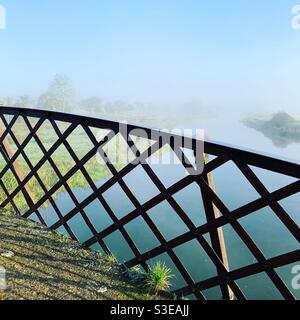 Una mattinata foggosa con il sole luminoso di un ponte pedonale in ferro battuto sul fiume Nene nel Northamptonshire, Inghilterra. Foto Stock