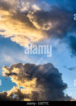 Raggi di luce solare che irradiano raggi di sole attraverso le nuvole bianche e grigie, cielo blu Foto Stock
