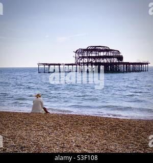 Donna seduta sulla spiaggia di Brighton alle rovine del vecchio West Pier Sussex, Regno Unito Foto Stock