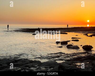 Due pescatori si sono sdraiati contro il sole che tramonta sulla costa del Galles del Sud, luglio. Foto Stock