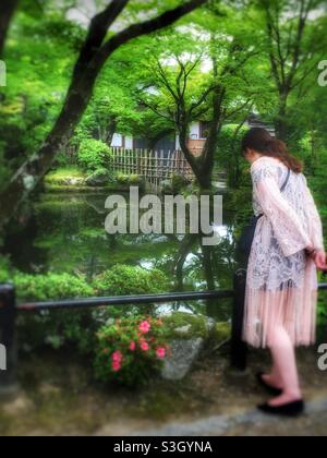 Ragazza che guarda in lago nei giardini del tempio Kiyomizu-dera, Kyoto, Giappone, con ponte, alberi e piccolo edificio bianco in background. Foto Stock