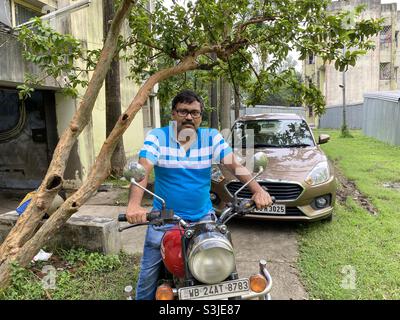 Uomo indiano di mezza età si siede su due ruote in una zona residenziale del Bengala con quattro ruote sullo sfondo Foto Stock
