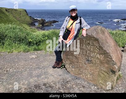 Una donna caucasica di 66 anni che posa contro un cartello di masso al Giants Causeway in Irlanda. Foto Stock