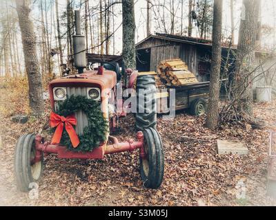 Foto vignettata di trattore rosso vintage con corona di Natale, carico di rimorchio di legno di fuoco, e capannone di legno Foto Stock