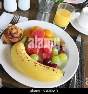 Una scelta di stile di vita sano per la prima colazione include frutta assortita e un rotolo di cannella servito con un bicchiere di succo d'arancia. Foto Stock