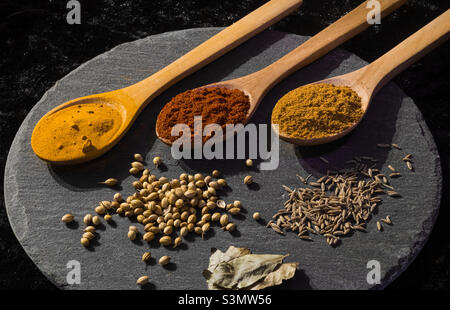 3 cucchiai di legno pieni di spezie, con semi e foglie su un asse di ardesia Foto Stock