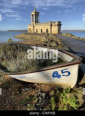 Una vista della chiesa di Normanton alla fine del suo accesso strada, per metà sommerso in acqua Rutland, con una piccola barca metà piena di vegetazione in primo piano Foto Stock