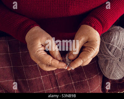 Vista dall'alto delle mani di una donna anziana, vestita con un maglione borgogna e coperta da una coperta a scacchi, si siede su una poltrona e si maglia con fili grigi. Foto Stock