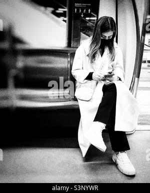 Una giovane donna in un elegante cappotto crema lungo seduto su un treno sotterraneo sta controllando il suo telefono cellulare. La foto è in bianco e nero. Foto Stock