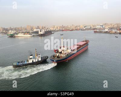 Piccola nave portarinfuse assistita da rimorchiatore durante l'approdo nel porto di Alessandria, Egitto. Sullo sfondo sono la costruzione della città e di altre navi ormeggiate durante il funzionamento del carico. Foto Stock