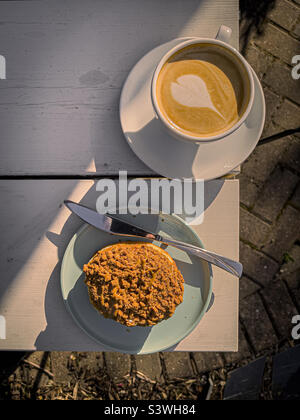 Tazza di caffè e un piatto con ciambella con cima adesiva sul bordo di un tavolo da giardino bianco. Foto Stock