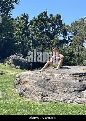 Una giovane donna siede su una roccia guardando il suo cellulare in una giornata estiva a Central Park, New York City, USA Foto Stock