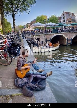 Un busker suona la sua chitarra seduto su un canale in una bella serata estiva durante l'ora d'oro nel cuore di Amsterdam Foto Stock