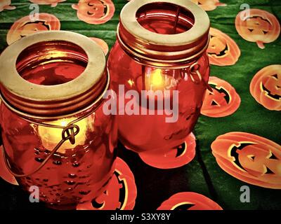 Un paio di candele giara di muratore arancione danno luce tremolante seduto su un tavolo coperto da una tovaglia stagionale di Halloween jack-o-lantern. Foto Stock