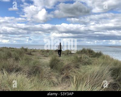 Un uomo si allontana dalla macchina fotografica, tra le erbe soffiate dal vento in un paesaggio costiero, con vista sul mare sullo sfondo e cielo blu con le nuvole sopra. Foto Stock