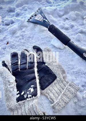 Un paio di guanti di lana e un attrezzo per neve/ghiaccio, un'estremità un raschietto per ghiaccio, l'altra estremità un pennello da neve. Una vita ancora ventosa si è imposta su un cofano di automobile coperto di neve dopo una tempesta di neve dello Utah, Stati Uniti. Foto Stock