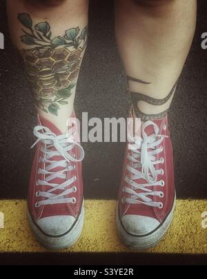 Guardando in basso le gambe tatuate di una giovane donna e le sneakers rosse alte Chuck Taylor. Foto Stock