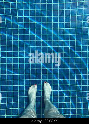 Guardando giù sui piedi e sulle gambe di una donna con le unghie dipinte di rosso in una piscina dalle piastrelle blu Foto Stock