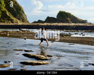 Un uomo porta una tavola da surf attraverso un piccolo fiume alla spiaggia di Samara Costa Rica Foto Stock