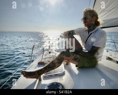 Un uomo con tatuaggi gode del sole dal ponte di un catamarano in mare calmo Foto Stock